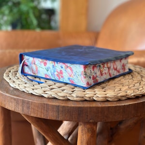 May include: A thick book with a solid dark blue cover and a decorative page edge featuring a colorful floral pattern of red, pink, and blue flowers on a white background. The book is closed and rests on a round, woven, light brown textured mat.