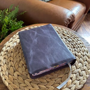 May include: A rectangular book with a dark purple, textured leather cover. The book features a white ribbon bookmark extending from the bottom edge, and the page edges display a colorful, intricate floral pattern with various shades of pink and blue.