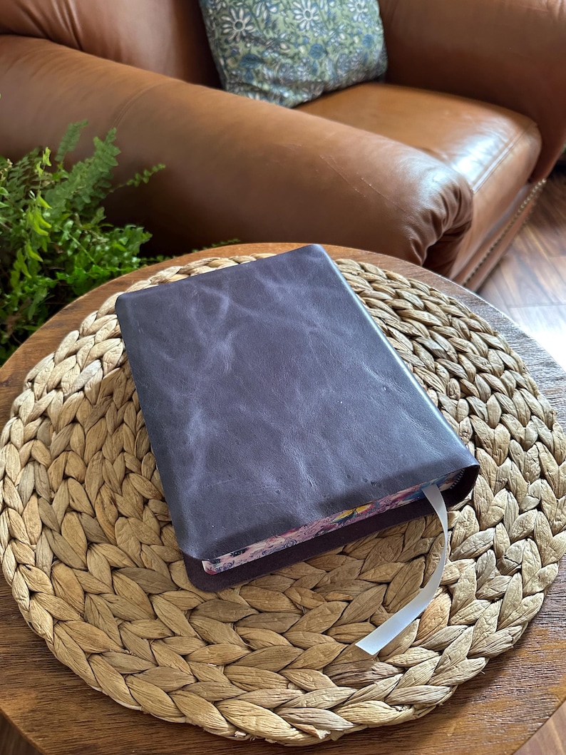 May include: A rectangular book with a dark purple, textured leather cover. The book features a white ribbon bookmark extending from the bottom edge, and the page edges display a colorful, intricate floral pattern with various shades of pink and blue.