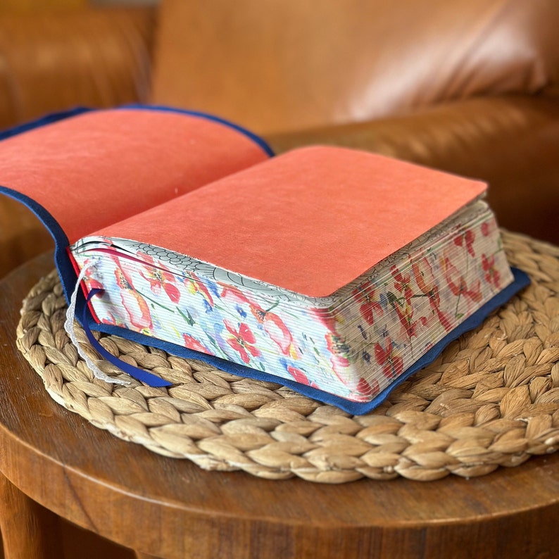 May include: A partially open book with a dark blue cover and a bright coral-colored endpaper. The page edges feature a vibrant floral pattern with red and pink flowers on a white background, accompanied by a thin white ribbon bookmark.