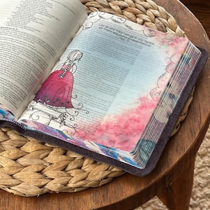 May include: An open book with a dark cover featuring a translucent vellum page overlay. The page displays a black ink illustration of a person in a dress with braided hair, alongside pink floral watercolor accents and the text Let all who take refuge in you rejoice, let them sing joyful praises forever.