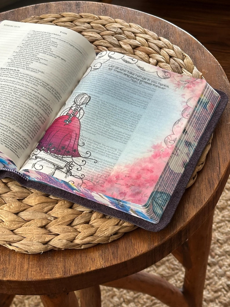 May include: An open book with a dark cover featuring a translucent vellum page overlay. The page displays a black ink illustration of a person in a dress with braided hair, alongside pink floral watercolor accents and the text Let all who take refuge in you rejoice, let them sing joyful praises forever.