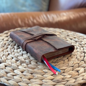 May include: A brown leather-bound journal with a strap closure and red, white, and blue ribbon bookmarks. The journal is sitting on a woven straw surface.