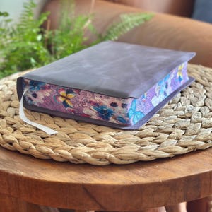 May include: A thick book with a plain dark gray leather cover and a white ribbon bookmark. The page edges feature a vibrant, colorful printed pattern of blue and yellow butterflies, pink flowers, and intricate swirling line designs.
