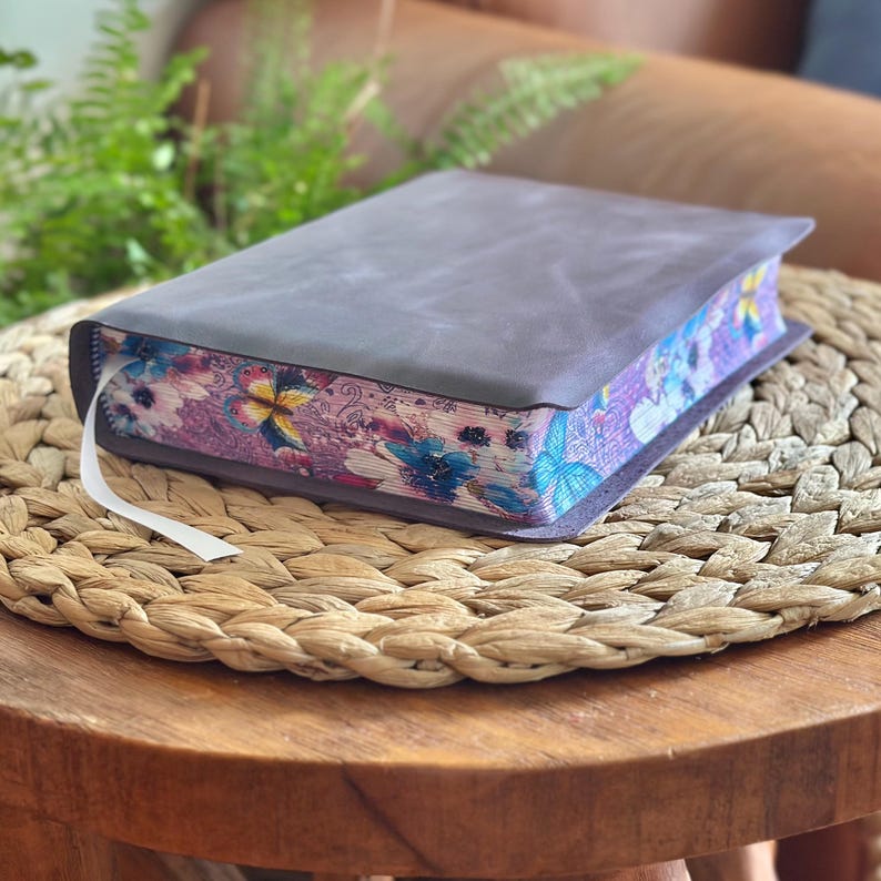 May include: A thick book with a plain dark gray leather cover and a white ribbon bookmark. The page edges feature a vibrant, colorful printed pattern of blue and yellow butterflies, pink flowers, and intricate swirling line designs.
