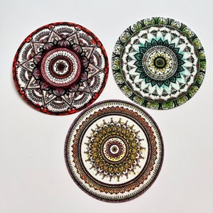 Könnte beinhalten: Drei runde Papieruntersetzer mit komplizierten Mandala-Designs. Die Untersetzer sind in verschiedenen Farben gehalten, darunter Rot, Grün, Gelb und Schwarz.