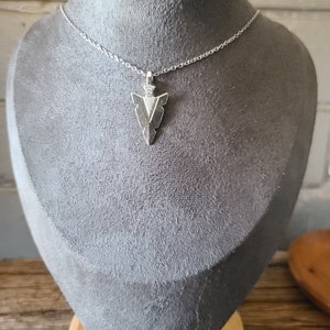 May include: A silver necklace with a silver arrowhead pendant. The necklace is on a grey mannequin.
