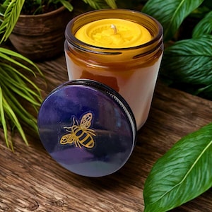 Puede incluir: Una vela en tarro de vidrio marrón con cera amarilla y una mecha blanca. La tapa azul oscuro presenta una ilustración de abeja dorada. La vela está sobre una superficie de madera, rodeada de hojas verdes.