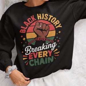 May include: Black sweatshirt featuring a circular design with the words "Black History" and a brown fist breaking chains. Below, the words "Breaking Every Chain" are displayed. The design incorporates red, yellow, and green.