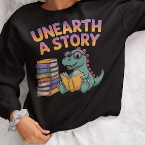 Może przedstawiać: Czarna bluza z napisem "UNEARTH A STORY" w kolorze fioletowym, pomarańczowym i żółtym. Kreskówkowy dinozaur w okularach czyta książkę obok stosu kolorowych książek. Motyw edukacyjny.