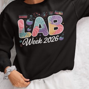 Może przedstawiać: Czarna bluza z napisem "LAB Week 2026" w kolorowym stylu tie-dye. Projekt zawiera ilustracje sprzętu laboratoryjnego i grafikę o tematyce naukowej. Bluza ma okrągły dekolt.