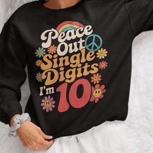 May include: Black sweatshirt with the text "Peace Out Single Digits I'm 10" in a retro font. The design includes a rainbow, peace sign, and flower graphics. The sweatshirt is a birthday-themed apparel item.
