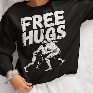 May include: Black sweatshirt with the words "FREE HUGS" in large white letters. Below the text is a graphic of two wrestlers embracing. The sweatshirt is a crew neck style.