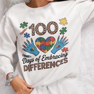 May include: White sweatshirt with a colorful graphic design. The design includes the number "100" in a leopard print pattern, puzzle pieces, a heart-shaped puzzle, and the text "Days of Embracing Differences."