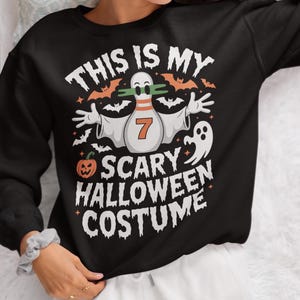 Op de afbeelding: Zwarte Halloween-trui met de tekst "THIS IS MY SCARY HALLOWEEN COSTUME". De afbeelding toont een bowlingpin-geest met nummer 7, vleermuizen, een pompoen en een spook.