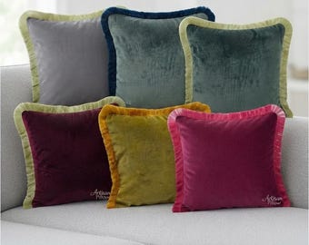 May include: Six square velvet throw pillows in gray, dark teal, green, deep red, mustard yellow, and magenta. Each pillow features a contrasting fringe trim along the edges and the text Artisan Pillow embroidered in the bottom corner of the covers.