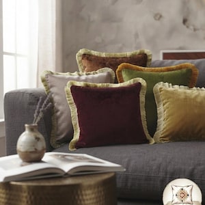 Pode incluir: Uma coleção de almofadas quadradas de veludo em várias cores, incluindo bordô, verde-oliva, amarelo-mostarda, marrom e cinza-claro. Cada almofada apresenta uma borda com franjas decorativas. O logotipo ARTISAN PILLOW é visível.