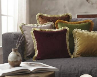 Puede incluir: Una colección de cojines cuadrados de terciopelo en varios colores, incluyendo burdeos, verde oliva, amarillo mostaza, marrón y gris claro. Cada cojín presenta un borde con flecos decorativos. El logotipo ARTISAN PILLOW es visible.