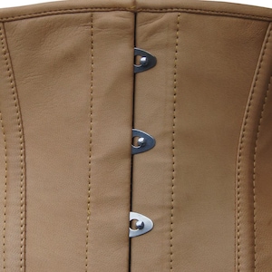 Op de afbeelding: Een beige leren korset met een verticale sluiting aan de voorzijde, voorzien van drie zichtbare zilverkleurige metalen gespen. Het kledingstuk is geconstrueerd met versterkte verticale naden en bijpassende beige stiksels over het gladde oppervlak.