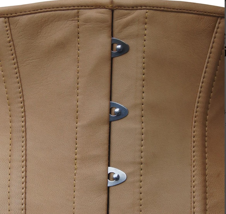 Op de afbeelding: Een beige leren korset met een verticale sluiting aan de voorzijde, voorzien van drie zichtbare zilverkleurige metalen gespen. Het kledingstuk is geconstrueerd met versterkte verticale naden en bijpassende beige stiksels over het gladde oppervlak.