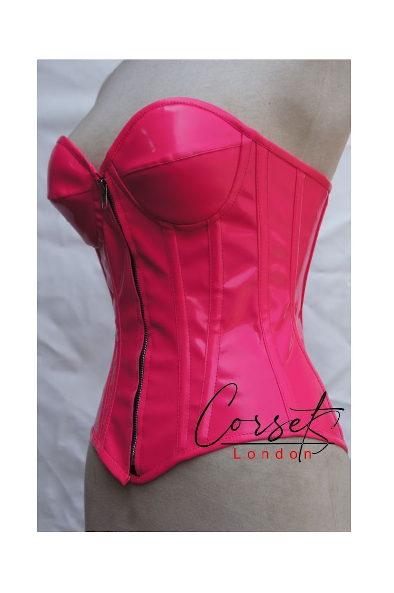 May include: A bright pink, strapless, vinyl corset with a front zip closure. The corset has boning and seams for structure. The text "Corset London" is printed on the bottom of the corset.