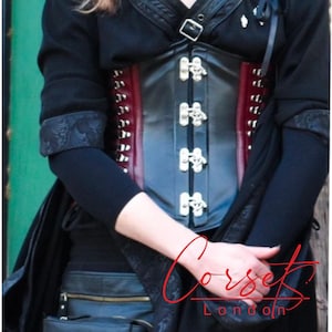 May include: A black leather corset with silver buckles and red lacing. The corset has a decorative design on the front and is worn by a person.