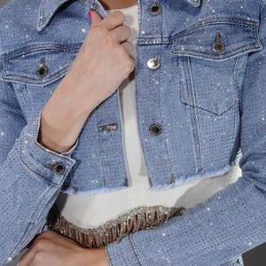 May include: Light blue denim jacket with rhinestone embellishments. The cropped jacket features a classic collar, button closures, and two front pockets. The jacket is paired with a matching belt and jeans.