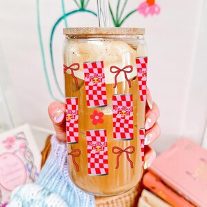 Könnte beinhalten: Ein durchsichtiges Glas mit Bambusdeckel und Glasstrohhalm, gefüllt mit Eiskaffee. Die Außenseite ist mit einem wiederkehrenden Muster aus rot-rosa karierten Energy-Drink-Dosen, roten Schleifen und kleinen roten Blumen bedruckt.