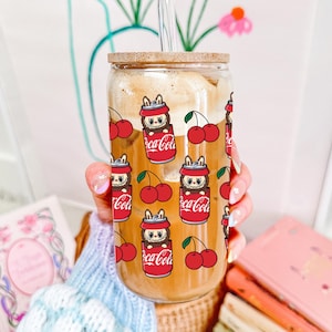 Könnte beinhalten: Ein durchsichtiges Glas mit Bambusdeckel und Glasstrohhalm, gefüllt mit Eiskaffee. Die Außenseite zeigt ein wiederkehrendes Muster aus Zeichentrickfiguren in roten Getränkedosen mit Coca-Cola-Aufschrift sowie leuchtend roten Kirschen.