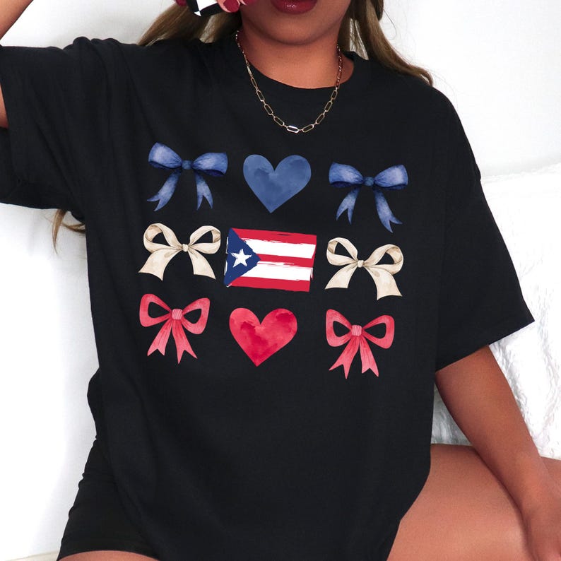 May include: Black t-shirt featuring a watercolor design of blue and cream bows, blue hearts, a red heart, and the Puerto Rican flag. The shirt is a casual, oversized fit.