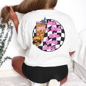 Op de afbeelding: Wit T-shirt met een afbeelding op de achterkant met een hamburger, drankje en de tekst "Just a girl who loves Sonic" in roze en zwart. De afbeelding is geplaatst tegen een zwart-witte geruite achtergrond.