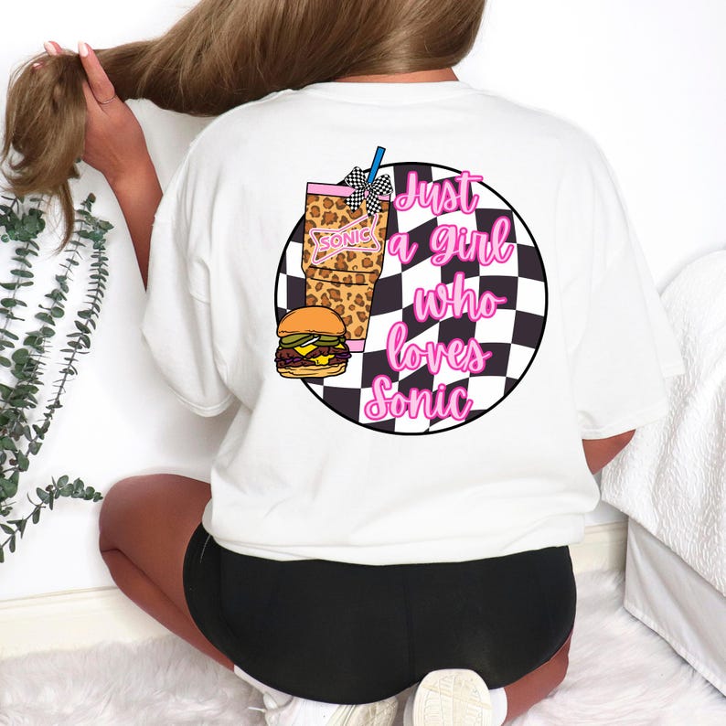 Op de afbeelding: Wit T-shirt met een afbeelding op de achterkant met een hamburger, drankje en de tekst "Just a girl who loves Sonic" in roze en zwart. De afbeelding is geplaatst tegen een zwart-witte geruite achtergrond.