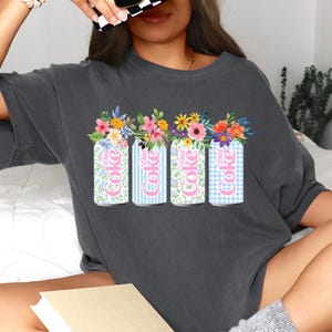 K&ouml;nnte beinhalten: Dunkelgraues T-Shirt mit vier Diet Coke-Dosen mit Blumenarrangements. Die Dosen haben verschiedene Muster: floral, blaue Streifen, floral und blaues Vichy-Karo. Die Worte "Diet Coke" sind auf jeder Dose zu sehen.