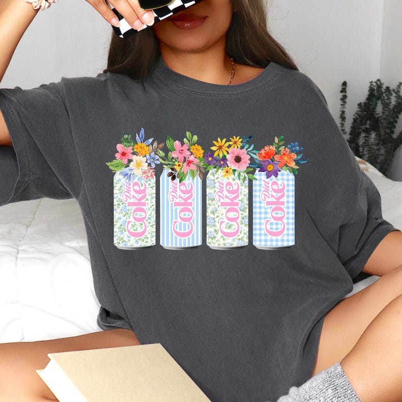 K&ouml;nnte beinhalten: Dunkelgraues T-Shirt mit vier Diet Coke-Dosen mit Blumenarrangements. Die Dosen haben verschiedene Muster: floral, blaue Streifen, floral und blaues Vichy-Karo. Die Worte "Diet Coke" sind auf jeder Dose zu sehen.