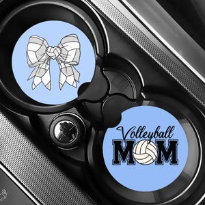Puede incluir: Dos posavasos para coche con fondos azules. Uno presenta un dise&ntilde;o de lazo de voleibol en blanco y gris. El otro dice "Volleyball MOM" con un gr&aacute;fico de voleibol en negro, blanco y beige.
