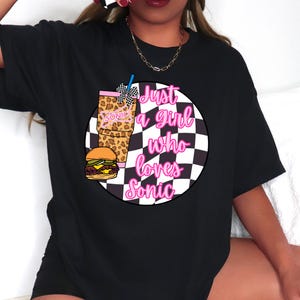 Op de afbeelding: Zwart T-shirt met een afbeelding van een Sonic-drankje, een hamburger en de tekst "Just a girl who loves Sonic" in roze letters. De drank heeft een luipaardprint en een geruite strik.