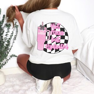 Puede incluir: Camiseta blanca con un gr&aacute;fico de vaso Stanley rosa brillante y el texto "Hot mess but Hydrated" en rosa. El vaso tiene un lazo a cuadros blanco y negro y est&aacute; colocado contra un c&iacute;rculo a cuadros blanco y negro.