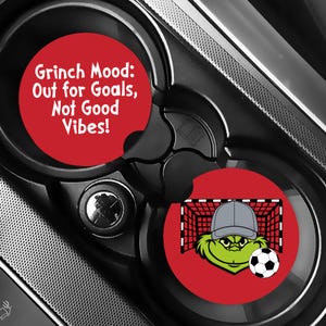 Puede incluir: Dos posavasos rojos para coche con dise&ntilde;os del Grinch. Uno tiene el texto "Grinch Mood: Out for Goals, Not Good Vibes!". El otro presenta al Grinch con una gorra gris, un bal&oacute;n de f&uacute;tbol y una porter&iacute;a.