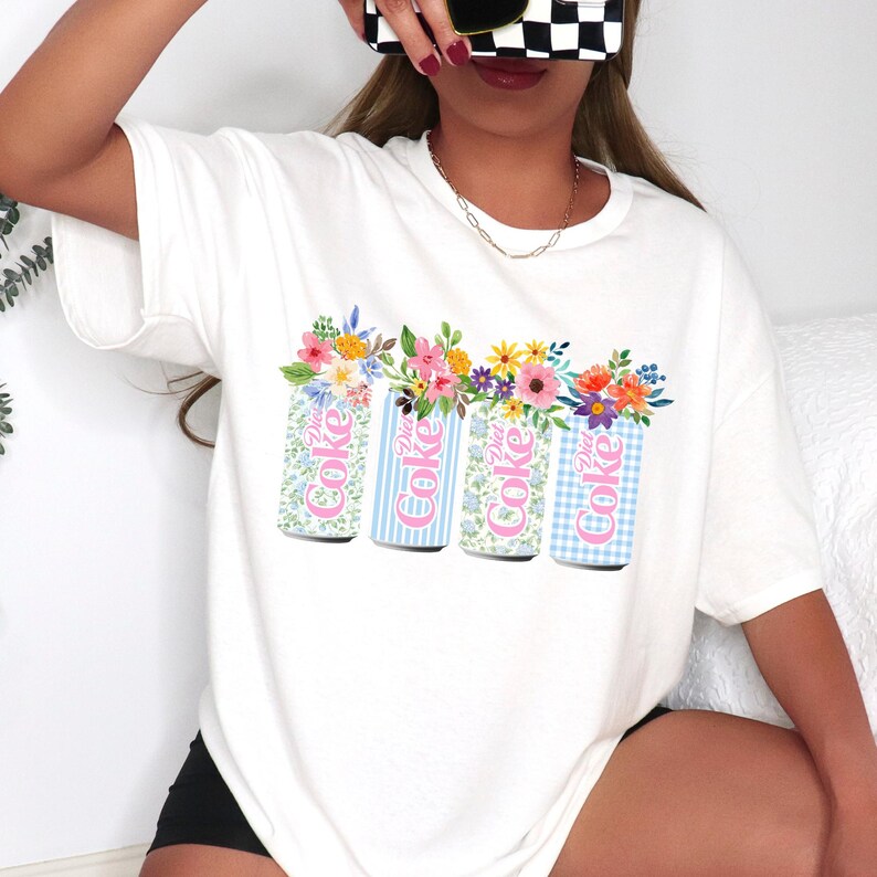 K&ouml;nnte beinhalten: Wei&szlig;es T-Shirt mit einer Grafik von vier Diet Coke-Dosen, die mit Blumen verziert sind. Die Dosen haben verschiedene Muster: floral, blaue Streifen und blaues Vichy. Der Text "Diet Coke" ist auf jeder Dose zu sehen.
