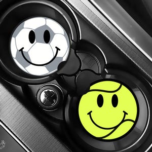 Puede incluir: Dos posavasos para coche con caras sonrientes. Uno es un dise&ntilde;o de bal&oacute;n de f&uacute;tbol con una cara sonriente. El otro es un dise&ntilde;o de pelota de tenis con una cara sonriente. Ambos posavasos est&aacute;n en una consola de coche negra.