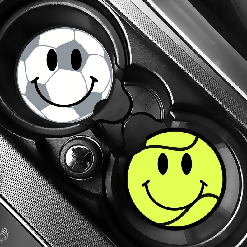 Puede incluir: Dos posavasos para coche con caras sonrientes. Uno es un dise&ntilde;o de bal&oacute;n de f&uacute;tbol con una cara sonriente. El otro es un dise&ntilde;o de pelota de tenis con una cara sonriente. Ambos posavasos est&aacute;n en una consola de coche negra.