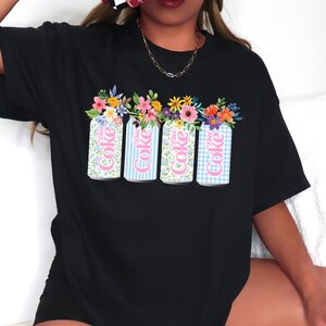 K&ouml;nnte beinhalten: Schwarzes T-Shirt mit einem Aufdruck von vier Diet Coke-Dosen mit Blumen. Die Dosen haben florale, blau gestreifte und karierte Muster. Die Worte "Diet Coke" sind in Rosa aufgedruckt.