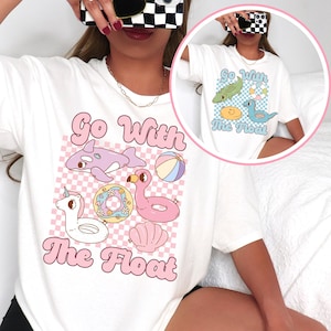 Könnte beinhalten: Weißes T-Shirt mit einem Grafikdesign mit dem Text "Go With The Float" und Illustrationen eines Wals, Flamingos, Einhorns, Donuts, Strandballs und einer Muschel. Das Design hat eine Retro- und verspielte Ästhetik.