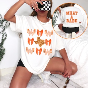 May include: White t-shirt featuring orange bow and "W" graphics, and a leopard print Texas shape. The back of the shirt has the text "WHAT A BABE" in orange. A casual, comfortable style.