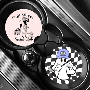 Puede incluir: Dos posavasos para coche con dise&ntilde;os de golf. Uno es de color rosa claro con una ilustraci&oacute;n vintage de una golfista y el texto "Golf Wives Social Club". El otro presenta un fantasma de dibujos animados con un palo de golf y un fondo a cuadros.
