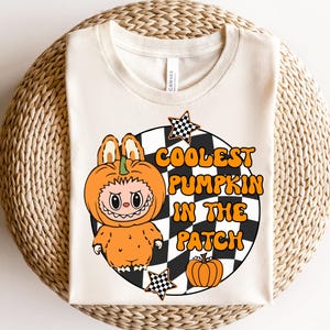 Könnte beinhalten: Cremefarbenes T-Shirt mit einem Cartoon-Charakter in einem orangefarbenen Kürbiskostüm. Das Design enthält den Text "COOLEST PUMPKIN IN THE PATCH" in Orange und Weiß, vor einem schwarz-weiß karierten Hintergrund mit Sternakzenten.