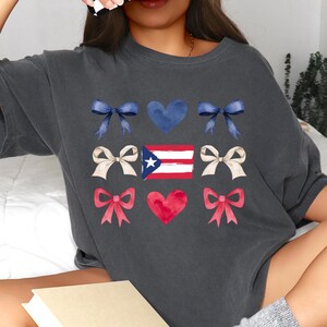 May include: Dark gray t-shirt featuring a design with blue bows, a blue heart, cream bows, a Puerto Rican flag, red bows, and a red heart. The design is a watercolor style. The shirt is a casual style.