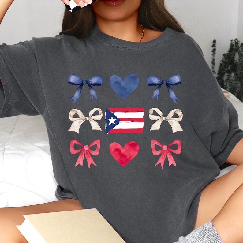 May include: Dark gray t-shirt featuring a design with blue bows, a blue heart, cream bows, a Puerto Rican flag, red bows, and a red heart. The design is a watercolor style. The shirt is a casual style.