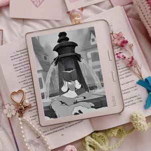Könnte beinhalten: Ein hellrosa Kobo-E-Reader mit einer schwarz-weißen Illustration einer Figur, die an einem Brunnen liest. Das Gerät hat seitliche Tasten und ist von einem Buch, einem Perlen-Schlüsselanhänger mit Herzanhänger und einer kleinen Figur umgeben.