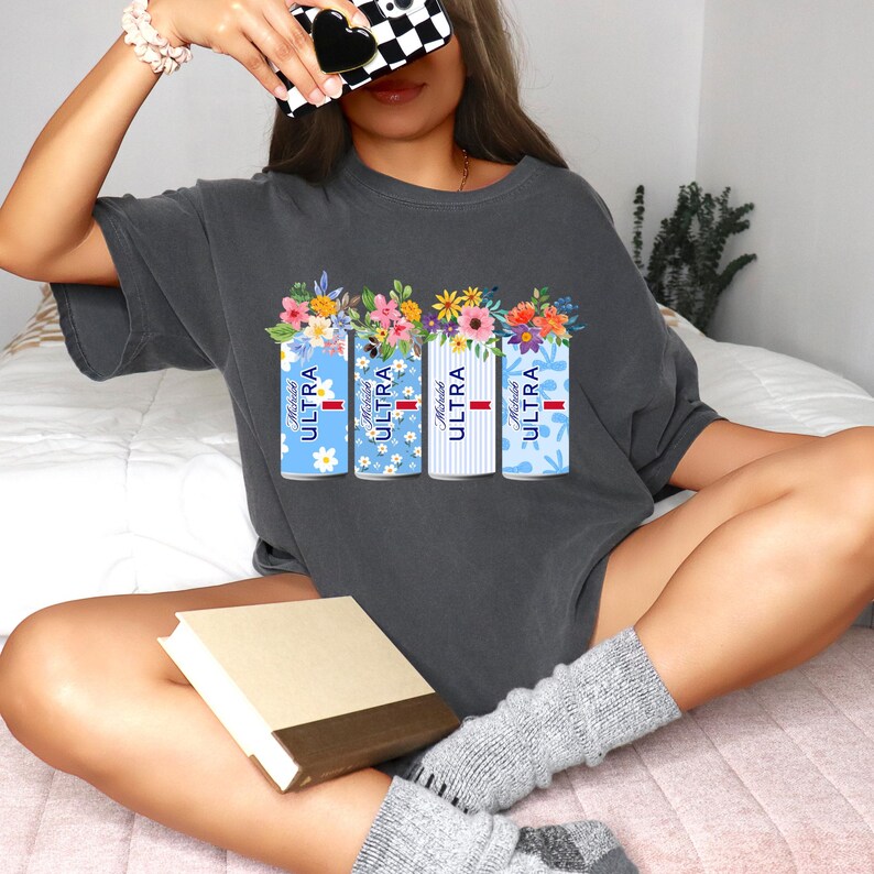 Puede incluir: Camiseta gris oscuro con cuatro latas de cerveza estilizadas con arreglos florales. Cada lata tiene un patr&oacute;n azul diferente y el texto "Michelob ULTRA". Las flores son amarillas, rosas y naranjas.